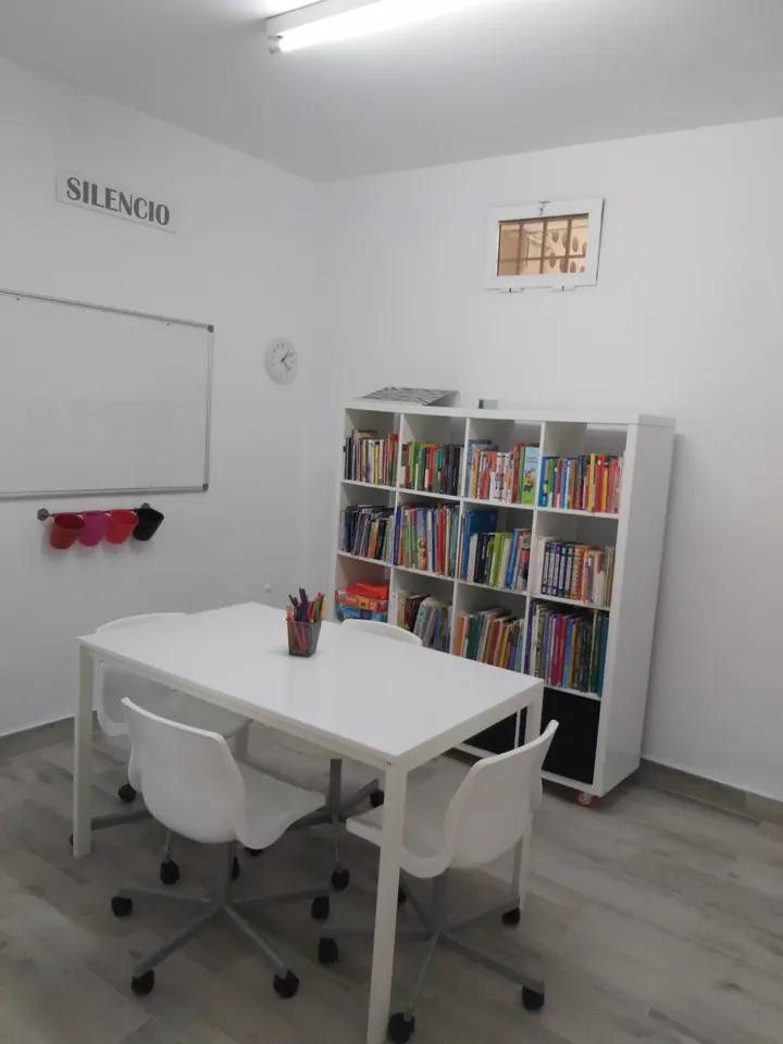 Aula pequeña con mesa, sillas blancas y estantería de libros